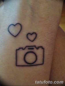 Фото тату небольшого фотоаппарата, окруженного двумя сердечками, выполненного тонкими линиями на руке.