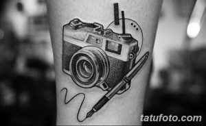 Фото тату: черно-белая татуировка старинного фотоаппарата и пера, выполненная в стиле олдскул на руке. Детализированный рисунок с тонкими линиями.