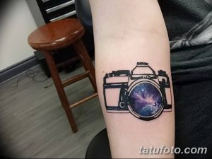 Фото тату: стилизованный фотоаппарат с изображением галактики в объективе, выполненный в черно-белой гамме с цветными вставками на предплечье.