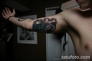 Черно-серая татуировка фотоаппарата на бицепсе мужчины. Реалистичный рисунок с детальной проработкой и четкими контурами.