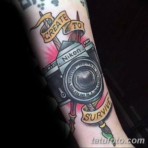 Фото тату фотоаппарата Nikon на предплечье, украшенного лентой с надписью 'CREATE TO SURVIVE' и брызгами краски. Яркий и символичный рисунок татуировки.