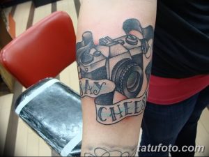 Фотография татуировки с изображением винтажного фотоаппарата и надписью "Say Cheese" на руке. Черно-серый рисунок в стиле олдскул.