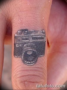Фотография маленькой татуировки фотоаппарата на пальце. Черно-серый рисунок, детализированный для своего размера.