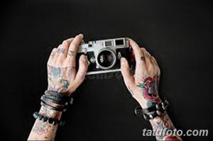 Фото рук, покрытых татуировками, держащих винтажный фотоаппарат. Рисунок татуировки на руках выполнен в различных стилях, включая традиционный и акварель.