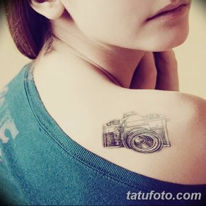 Фото тату небольшого, схематичного фотоаппарата на лопатке девушки. Чёрно-белый рисунок, выполненный тонкими линиями, выглядит элегантно и стильно.