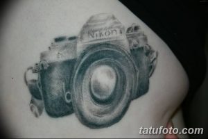 Черно-серая татуировка камеры Nikon на плече. Рисунок выполнен в графическом стиле с использованием теней и полутонов.