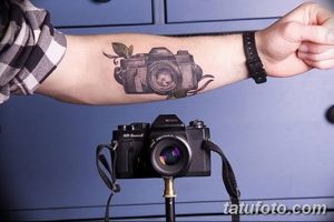 Фото татуировки фотоаппарата на руке, украшенного красными брызгами краски. Реалистичный рисунок тату для любителей фотографии.