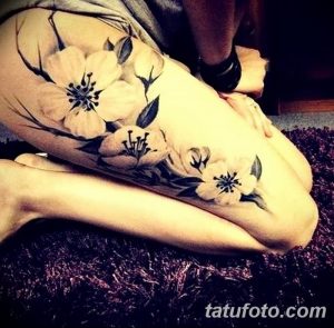 Фотография татуировки на бедре, изображающей цветущую ветку сакуры. Рисунок выполнен в нежных пастельных тонах с использованием черных контуров.