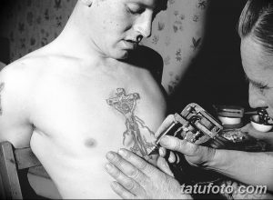 Фотография процесса нанесения татуировки в стиле олд скул, сделанная в черно-белом цвете. Виден мастер, работающий машиной над татуировкой на груди клиента.