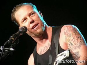 Фотография Джеймса Хетфилда из Metallica, выступающего на сцене с микрофоном. Видны татуировки на его руках и плечах.