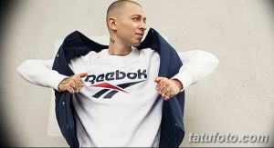Фото мужчины с татуировками на руках, демонстрирующего логотип Reebok на своей одежде. Татуировки на костяшках пальцев с надписью 'IMPERIVM' и на предплечье.