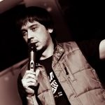 Фото тату на руке комика, выступающего с микрофоном. Видны детали рисунка татуировки, выполненной в темных тонах.