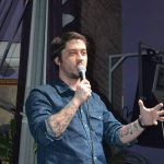 Фото мужчины с татуировкой на руке, выступающего с речью на сцене. Видны детали татуировки на предплечье.