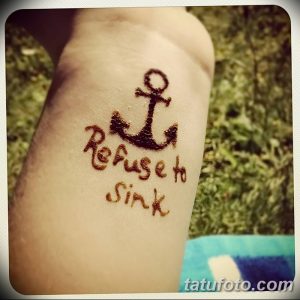 Фото тату якоря с надписью 'Refuse to sink' на предплечье. Рисунок выполнен в минималистичном стиле, коричневыми чернилами.