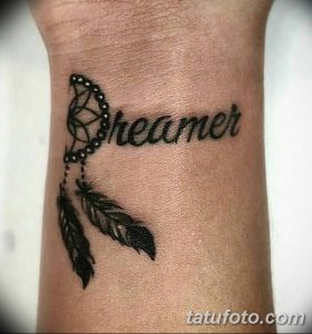 Татуировка на запястье, изображающая слово "Dreamer" (Мечтатель) с изображением ловца снов и двух перьев.