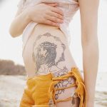Фото тату: изображение лица Иисуса Христа с терновым венцом на животе девушки, выполненное в графическом стиле.