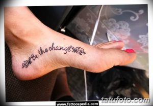 Фото тату: Черная надпись "Be the change" с небольшими цветочными элементами, выполненная на внешней стороне женской стопы. Минималистичный рисунок татуировки.