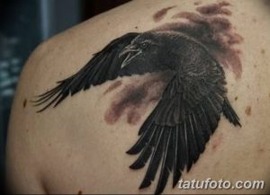 Фото реалистичной татуировки ворона на плече, окруженного облаками. Черно-серый рисунок с детализированными перьями.