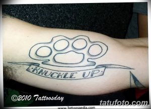 Фото тату: черно-белый контурный рисунок кастета с надписью "Knuckle Up" на руке, выполненный в минималистичном стиле.