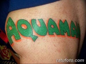 Фото тату на руке, изображающее надпись "AQUAMAN" зелеными буквами с оранжевой обводкой, выполненное в простом, но эффектном стиле.