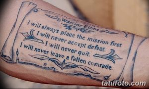 Фото тату: реалистичное изображение свитка с текстом "Warrior Ethos" на предплечье, выполненное в черно-серых тонах.