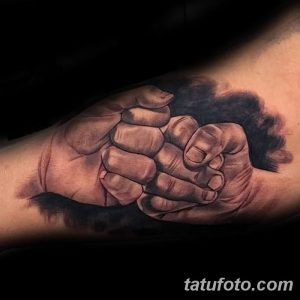 Фотография реалистичной татуировки с изображением сцепленных мужских рук на предплечье. Тату выполнено в черно-серых тонах с детальной проработкой кожи и мышц.
