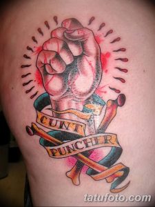 Фото татуировки на руке: сжатый кулак с надписью 'CUNT PUNCHER' на ленте и костями.