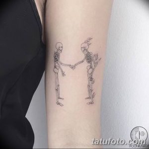 Фото тату: два скелета, держащиеся за руки и танцующие, на предплечье. Черно-белый рисунок татуировки с детальной прорисовкой костей и легким эффектом дымки.
