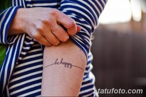 Черно-белое фото татуировки на запястье с надписью 'be happy', выполненной изящным рукописным шрифтом и соединенной с тонкой линией.