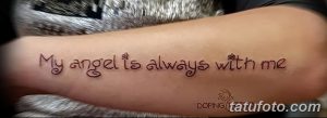 Фото тату: надпись 'My angel is always with me' и изображение маленького цветка на предплечье девушки. Красные и черные чернила.
