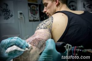 Фото тату: мастер наносит полинезийский орнамент на руку клиента. Виден процесс работы, тату-машинка и эскиз на коже.