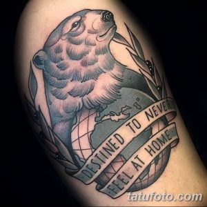 Фото тату медведя, стоящего на глобусе, с надписью 'Destined to never feel at home'. Рисунок выполнен в традиционном стиле с использованием черных и серых оттенков.
