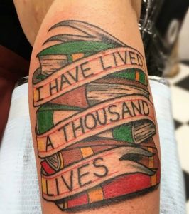 Фото татуировки на руке с изображением стопки книг и надписи "I Have Lived A Thousand Lives". Стиль олдскул, яркие цвета.