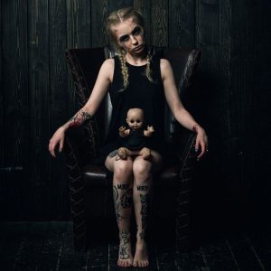 Фотография девушки с татуировками на руках и ногах, сидящей на кресле с куклой на руках.
