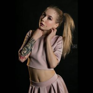 Фотография девушки в розовом наряде с татуировкой на руке, позирующей на фоне черного фона.