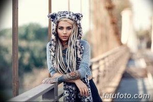Фотография девушки с татуировками на руках и шее, одетой в капюшон с ушками, на фоне моста, демонстрирующая загадочный и привлекательный образ.