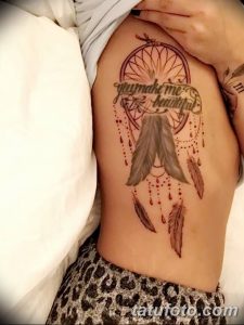 Фото тату: большой ловец снов на боку девушки, с перьями и надписью 'you make me beautiful'. Эскиз татуировки выполнен в коричневых тонах.