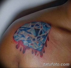 Фото тату бриллианта на плече. Изображение выполнено в акварельной технике с использованием голубых и красных оттенков. Капли стекают вниз, создавая эффект таяния.