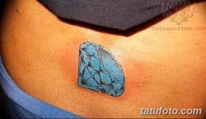 Фото тату небольшого голубого бриллианта на женской лопатке. Минималистичный рисунок татуировки выполнен в ярких тонах с четкими контурами.