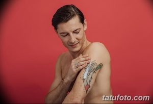 Фото татуировки на плече мужчины: кинжал, обвитый розой, выполненный в стиле олд скул. Мужчина трогает татуировку рукой.