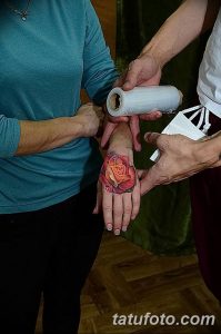 Фото свежей татуировки в виде красной розы на руке, обернутой защитной пленкой. Видны детали рисунка и процесс ухода за татуировкой.