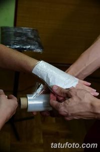 Фото процесса обертывания свежей татуировки на руке прозрачной защитной пленкой. Мастер тату аккуратно наносит пленку, обеспечивая гигиеническую защиту рисунка.