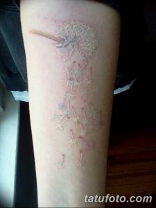 Фото тату одуванчика на ноге, выполненного в технике white ink. Изображение состоит из пушистых семян, разлетающихся по ветру.