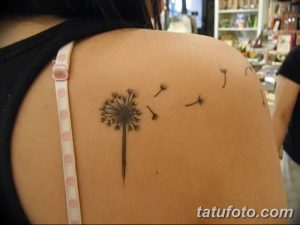 Фото тату одуванчика на плече. Небольшой, изящный рисунок с летящими парашютиками, выполненный тонкими линиями в черном цвете.