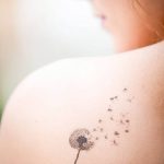 Фото минималистичной татуировки одуванчика на лопатке. Черно-белый рисунок татуировки, выполненный в тонких линиях, символизирующий легкость и свободу.