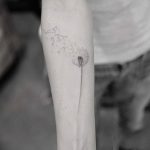 Фото тату: Черно-белый минималистичный рисунок одуванчика с летящими семенами, расположенный на предплечье. Эскиз татуировки выполнен тонкими линиями.