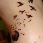 Фото татуировки с изображением одуванчика, из которого вылетает стая птиц, выполненной в черных тонах на плече.