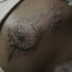 Фото татуировки одуванчика на лопатке. Черно-серый рисунок с разлетающимися семенами, выполненный в минималистичном стиле.