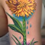 Фото реалистичной татуировки одуванчика на плече. Яркий рисунок татуировки с использованием желтых, зеленых и синих оттенков, выполненный опытным мастером тату.