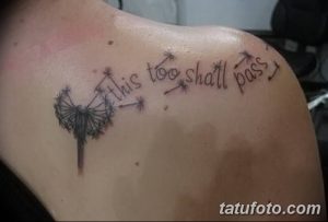 Фото тату одуванчика с надписью "This too shall pass" на плече. Черно-белый рисунок татуировки выполнен в минималистичном стиле с акцентом на смысл.